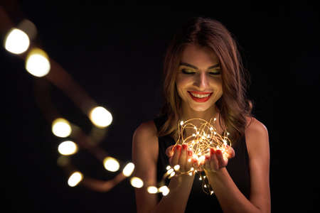 Party, holidays and celebration concept. Closeup of smiling woman holding Christmas light garland in palms, looking at camera, over dark background with bokehの写真素材