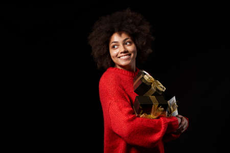 Black Friday sale, gift, present, celebration concept. Happy afro-american female holding a heap of black gift boxes with golden bows over black background, looking at blank copy spaceの写真素材