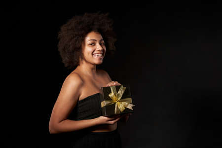 Black Friday sale, gift, present, celebration concept. Happy afro-american female holding one black gift box with golden bow over black background, looking at cameraの写真素材