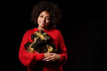 Black Friday sale, gift, present, celebration concept. Smiling afro-american female holding a heap of black gift boxes with golden bows over black background, looking at cameraの写真素材
