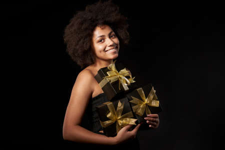 Black Friday sale, gift, present, celebration concept. Happy afro-american female holding many black gift boxes with golden bows over black background, looking at cameraの写真素材