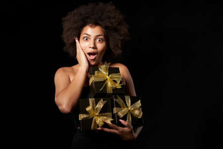 Black Friday sale, gift, present, celebration concept. Happy excited surprised afro-american female holding many black gift boxes with golden bows over black background, screaming of joyの写真素材