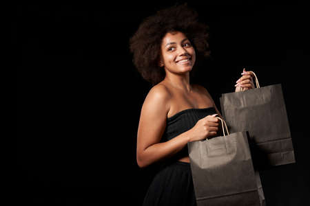 Black Friday sale, gift, present, celebration concept. Smiling afro-american female holding black paper shopping bags over black background, looking to side at blank copy spaceの写真素材