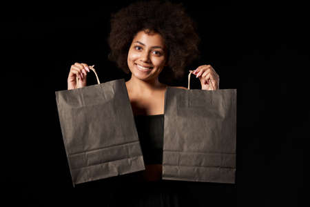 Black Friday sale, gift, present, celebration concept. Smiling afro-american female showing black paper shopping bags over black backgroundの写真素材