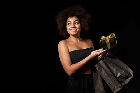 Black Friday sale, gift, present, celebration concept. Smiling afro-american female holding black paper shopping bags and a gift box, over black background, looking to side at blank copy spaceの写真素材