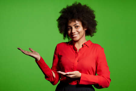 Smiling young African-American girl looking at camera pointing open hand palm at blank copy space, over green backgroundの写真素材
