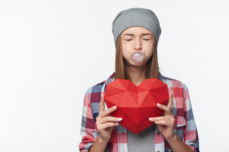 Funny teen girl holding red polygonal paper heart shape blowing bubblegum screwing up her eyes, studio portraitの写真素材