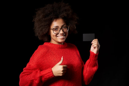 Happy young African-American girl in glasses and red sweater holding blank credit card and gesturing thumb up, over black backgroundの写真素材