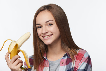 Closeup portrait of a teen girl holding half peeled banana going to eat it, looking at camera smilingの写真素材