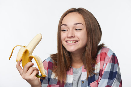 Closeup of happy teen girl holding half peeled banana going to eat it, looking at itの写真素材