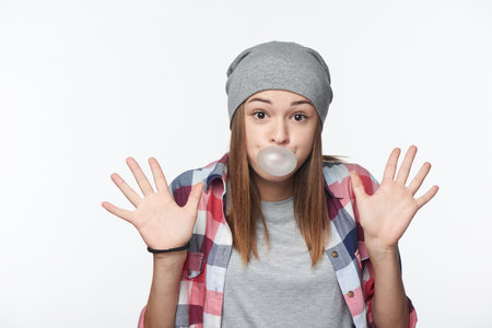 Surprised teen girl blowing bubblegum with arms raised, showing palms, studio portrait over white backgroundの写真素材