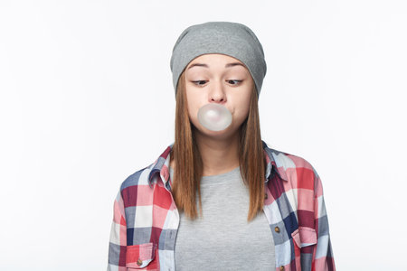 Funny portrait of teen girl blowing bubblegum and looking squinting at bubble, studio portrait over white background, isolatedの写真素材