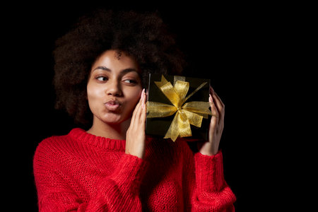 Black Friday sale, holidays gift, present, celebration concept. Closeup of happy afro-american female holding one black gift box with golden bow kissing it, over black background.の写真素材