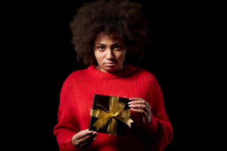Black Friday sale, gift, present, celebration concept. Discontent unhappy afro-american female holding a heap of black gift boxes with golden bows over black background, looking at camera broodingの写真素材