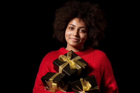 Black Friday sale, gift, present, celebration concept. Smiling afro-american female holding a heap of black gift boxes with golden bows over black background, looking at cameraの写真素材