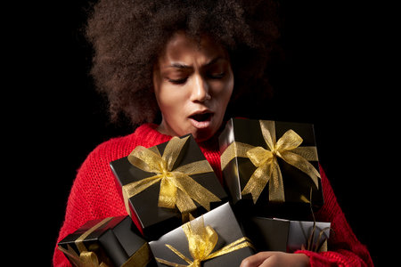Black Friday sale, gift, present, celebration concept. Emotional afro-american female holding a heap of black gift boxes with golden bows over black background, shoutingの写真素材