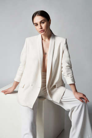 Fashion portrait of beautiful brunette woman in white jacket sitting on white box, studio shotの写真素材