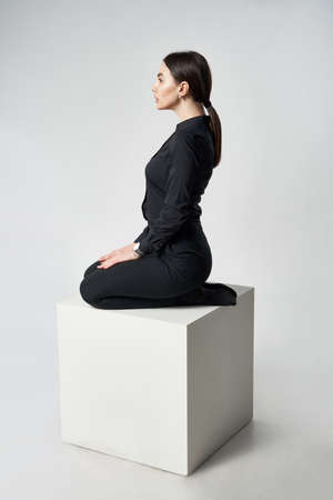 Stylish woman in black shirt and pants sitting on white box, side view portraitの写真素材