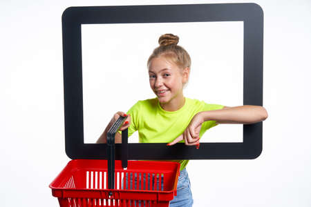 Online shopping concept. Trendy teen girl looking through digital screen holding empty shopping basket, pointing at it with surprise, isolated on light backgroundの写真素材