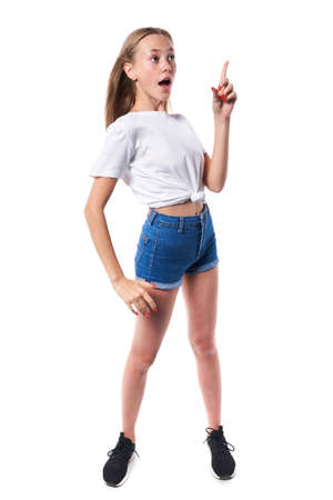 Full length of surprised teen girl wearing white t-shirt and denim shorts pointing up at blank copy space, over white backgroundの写真素材