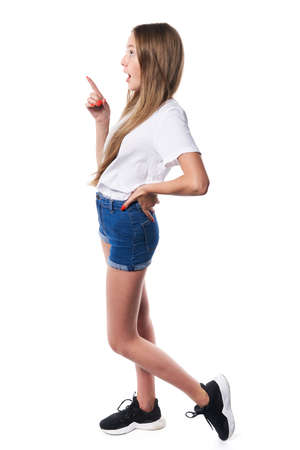 Full length profile of surprised teen girl wearing white t-shirt and denim shorts pointing to side at blank copy space, over white backgroundの写真素材