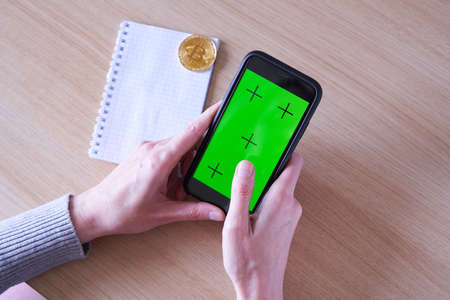 Trading app concept. Human hands cropped with smart phone with green screen, golden bitcoin coin and a notebookの写真素材