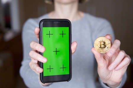 Cropped image of woman's hand showing green screen on smart phone and golden bitcoin coinの写真素材