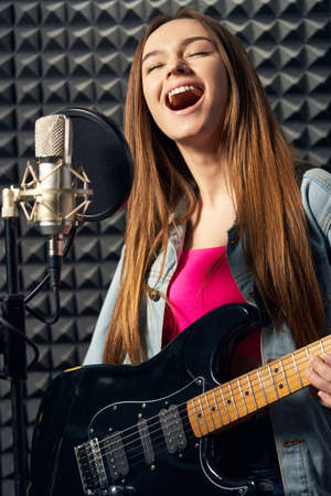 Girl musician in recording studio playing electric guitar and singing in micの写真素材
