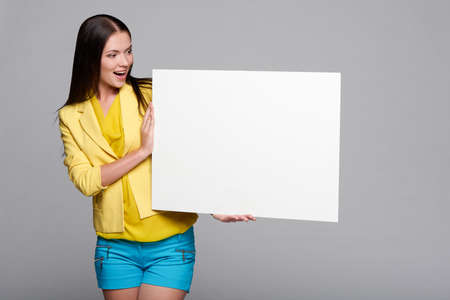 Surprised girl in yellow and blue, holding empty whiteboard, looking at blank copy space at boardの写真素材