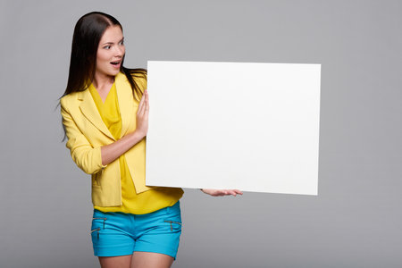 Surprised girl in yellow and blue, holding empty whiteboard, looking at blank copy space at boardの写真素材