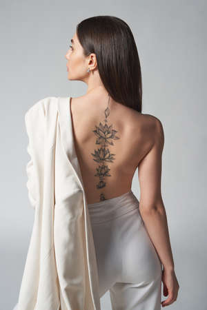 Back view fashion portrait of beautiful brunette woman holding white jacket over shoulder with tattoo on her backの写真素材