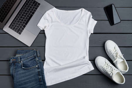 Flat lay of white t-shirt lying on rough wooden planks with laptop, smartphone and apparel accessories.の写真素材