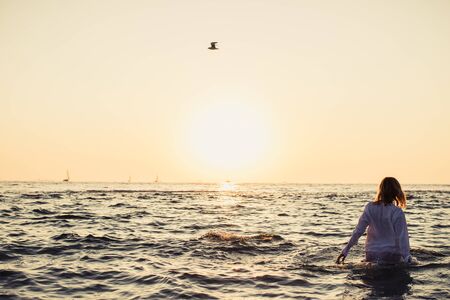 Woman in summer dress standing on a sea coast and looking to the horizon on sunrise. View backの写真素材