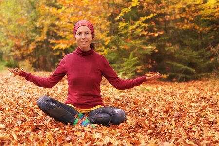 Girl in autumn sits on grass and cheerfully casts up yellow leaves in forest. Life style. Beautiful girl in colorful forest foliage outdoor.の写真素材
