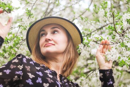 Beautiful  girl in straw hat sniffs blossom branch.  Portrait of a beautiful young blonde woman with long hair in a blue dress on  background of white cherry blossoms in spring. Spring season concept.の写真素材