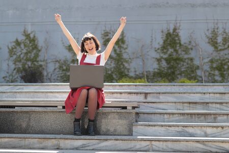 Girl rejoices in winning having raised her hands up sitting on steps in front of laptop. Technology, education and remote working concept, emotions of joyの写真素材