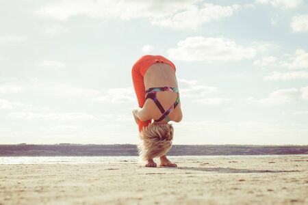 Young blond woman, wearing red leggings doing morning exercises on coast  in summer.