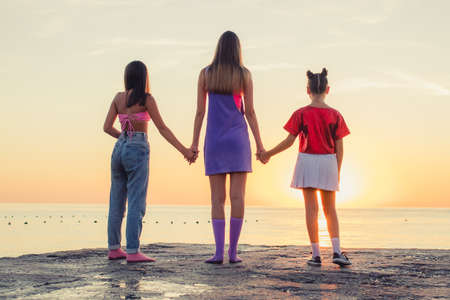 Back view of three young female friends standing on the beach holding hands and looking at the sea on sunrise.の写真素材