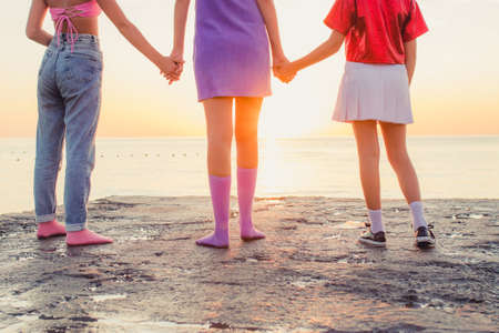Back view of three young female friends standing on the beach holding hands and looking at the sea on sunrise.の写真素材