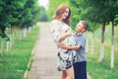 Cute pregnant woman in lace tunic hugs her son in the summer parkの写真素材