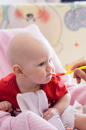 blonde baby in a red t-shirt tries first complementary foods from a small red spoon on a pink backgroundの写真素材