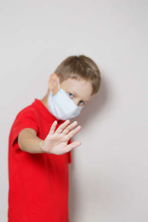 teenager with blond hair in a red T-shirt and a medical mask gets scared and repels the virus with his handの写真素材