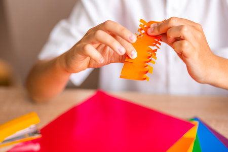 Boy's hands cut orange origami with orange scissors on multicolored paper background close-upの写真素材