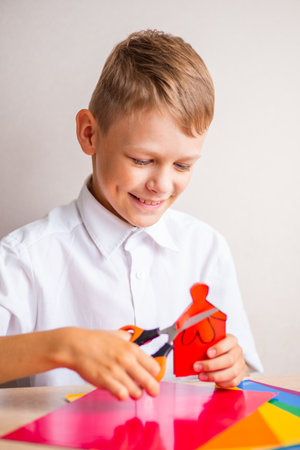 A smiling blond boy in a white cotton shirt is cutting a little man out of red paper with orange scissorsの写真素材