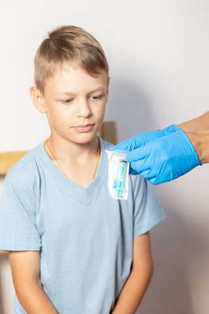 hands of a nurse in blue gloves open a pack of syringe close-up in front of the face of a boy in a blue t-shirtの写真素材