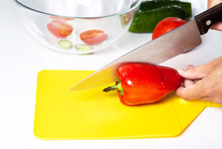 Female hands with a knife cut red pepper on a yellow cutting board.
