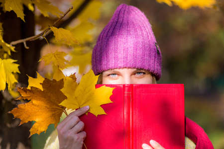 A girl in a knitted hat holds a maple leaf, covers her face with a book