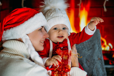 Mom in a Christmas hat shows daughter in a red outfit festive decorations on the background of the fireplaceの写真素材