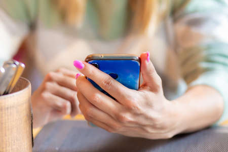Manicured female hands holding a smartphone while sitting at a table on a blurred coffee background.の写真素材