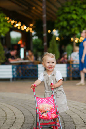 a little girl who has recently learned to walk is pushing a small toy stroller against a blurred background.の写真素材